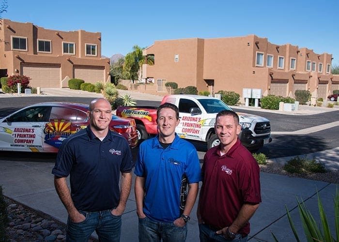 Arizona Painting Company's owners, with joyful smiles, leading the way in HOA painting services. Transforming community properties with quality coatings, boosting curb appeal and homeowner satisfaction. Three gentlemen in the image, one wearing black shirt on the left, in the middle a blue shirt and on the right a burgundy red shirt.